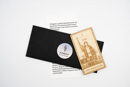 A tarot card set featuring a wooden 'The Magician' card with engraved details, displayed next to a black envelope with a circular 'MWM' logo sticker and printed cards on a white background.