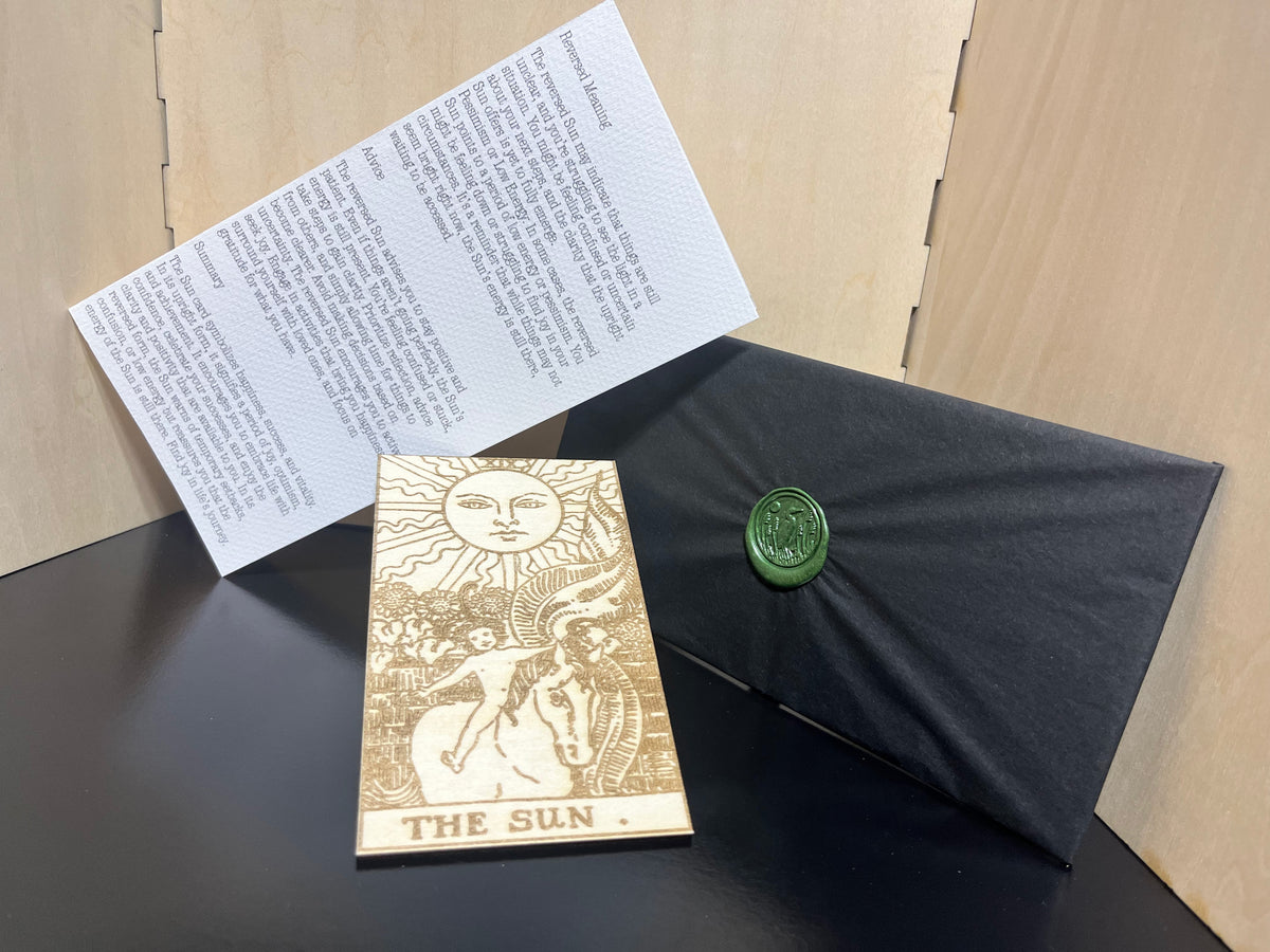 The image shows a wrapped and sealed black package with a gold-colored tarot card and its accompanying instruction sheet, labeled 'The Sun'.