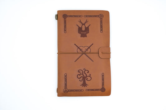 A brown leather journal with embossed Celtic-style designs, including a deer head, an oak tree, and an oak and knot symbol, secured with a leather wrap strap on a white background.
