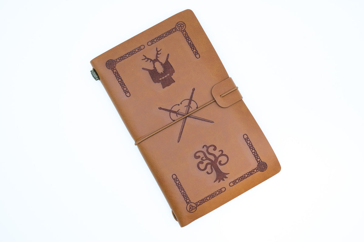 A tan leather journal with dark brown engraved designs, including a deer head, a wand and cauldron, and a tree, shown on a plain white background.