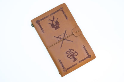 A tan leather journal with dark brown engraved designs, including a deer head, a wand and cauldron, and a tree, shown on a plain white background.