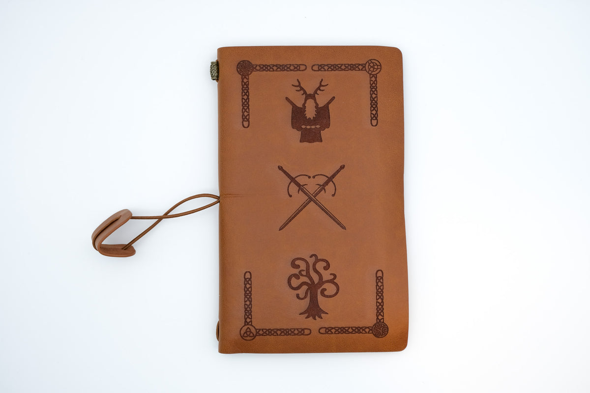 A brown leather journal with embossed Norse-inspired symbols and a matching leather wrist strap, shown on a clean white background.