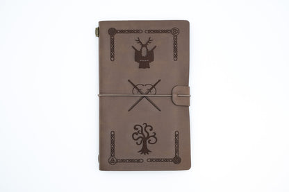 A brown leather journal with engraved black designs, including a stag, a tree, and crossed symbols, secured with a leather wrap closure on a white background.