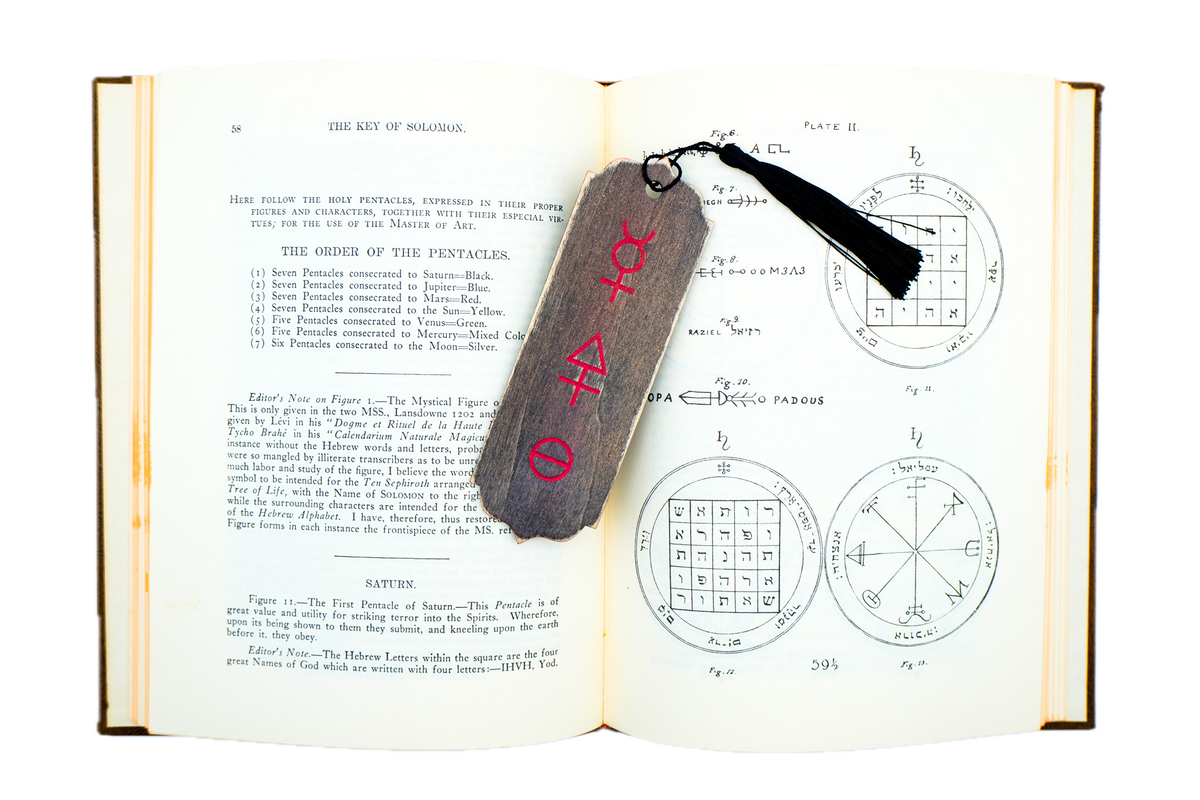 An open alchemical book showing printed pages with text and geometric diagrams, featuring a dark wooden tag with red tria prima alchemical symbols and a black tassel attached near the top right.