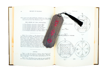 An open alchemical book showing printed pages with text and geometric diagrams, featuring a dark wooden tag with red tria prima alchemical symbols and a black tassel attached near the top right.