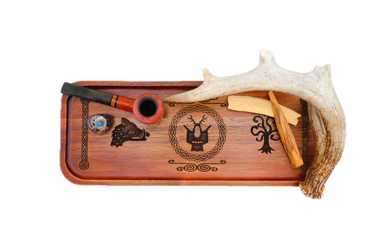 A rectangular wooden serving tray with a warm reddish-brown finish, featuring dark engraved symbols: a boar's head on the left, Cernunnos medallion, tree of life on the right, flanked by knotted pillar borders holding a reddish-brown smoking pipe, a small marbled glass bowl, and a curved antler from a top view.
