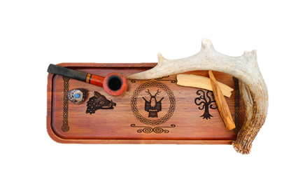 A rectangular wooden serving tray with a warm reddish-brown finish, featuring dark engraved symbols: a boar's head on the left, Cernunnos medallion, tree of life on the right, flanked by knotted pillar borders holding a reddish-brown smoking pipe, a small marbled glass bowl, and a curved antler from a top view.