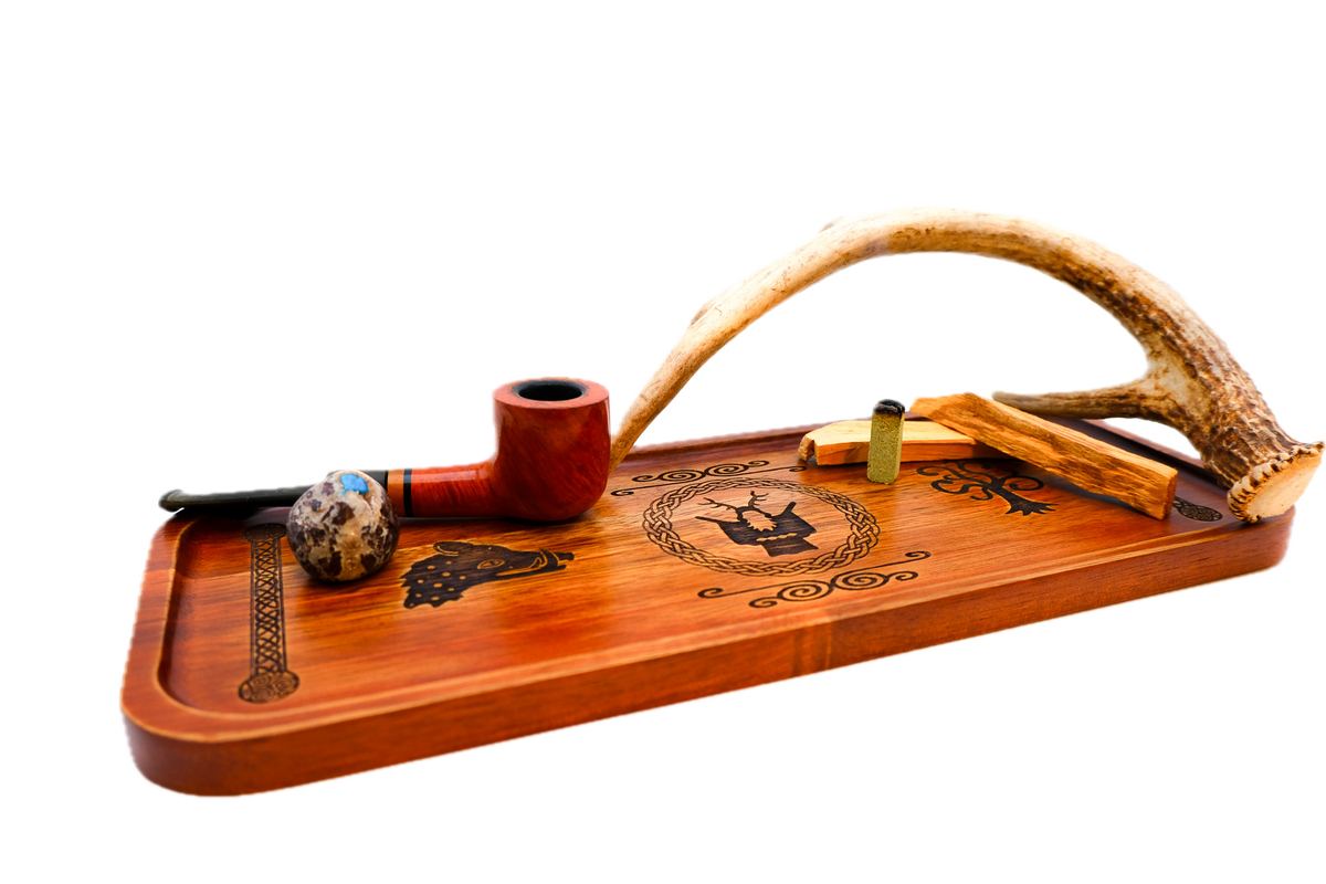 A rectangular wooden serving tray with a warm reddish-brown finish, featuring dark engraved symbols: a boar's head on the left, Cernunnos medallion, tree of life on the right, flanked by knotted pillar borders holding a reddish-brown smoking pipe, a small marbled glass bowl, and a curved antler.